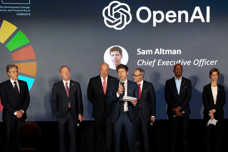 Un grupo de personas de pie en un escenario, vestidas con atuendo formal, con una persona sosteniendo un micrófono y hablando. El fondo muestra la "Abierto AI" logotipo, elementos gráficos coloridos y texto identificativo "Sam Altman, director ejecutivo." El escenario sugiere un evento o presentación formal.