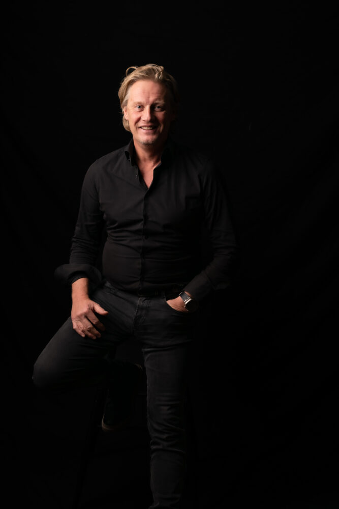 Een person en een zwart juvoly shirt en -broek zit op een kruk tegen een donkere achtergrond, glimlachend en vooruitkijkend, se reunió con la mano en zijn zak.