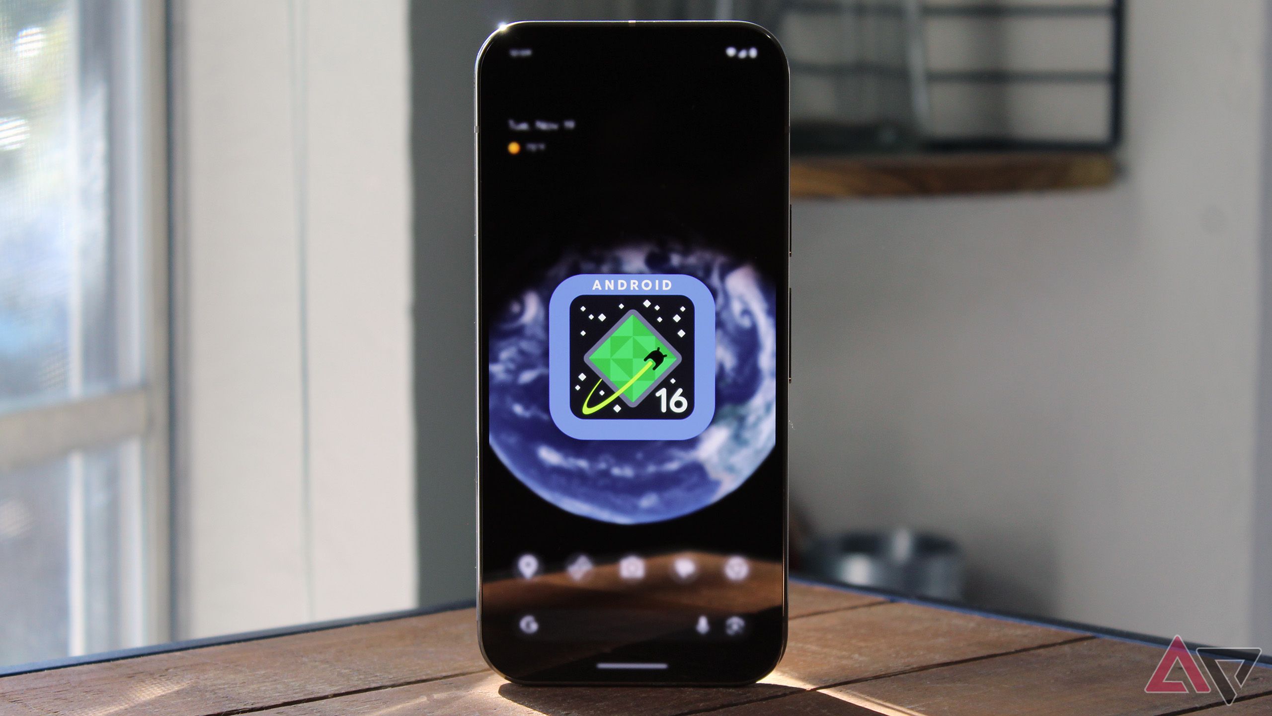Imagen del logotipo de Android 16 en la pantalla de inicio de Pixel 9 Pro con el teléfono sobre una mesa de madera.